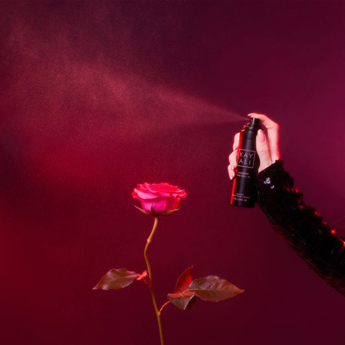 Hand spraying a black spray bottle labeled 'KAYALI' onto a red rose against a dark red background