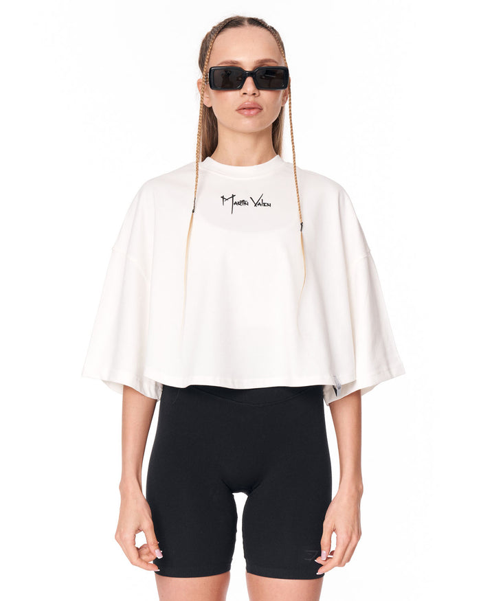 A model wearing a white cropped t-shirt with a black script logo on the chest, paired with black bike shorts and black sunglasses, against a plain white background.