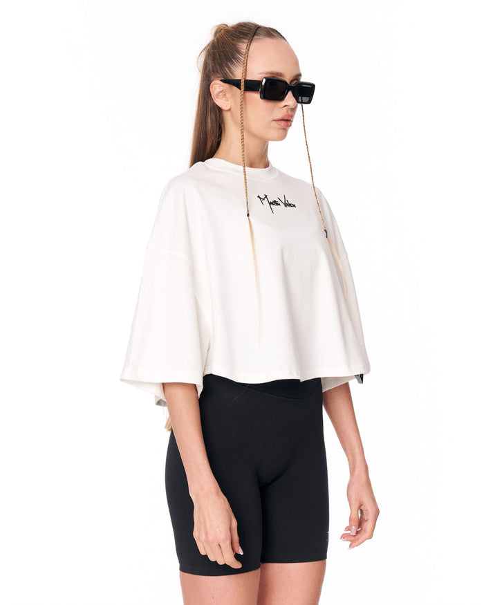 A model wearing a white cropped tunic top with a black script logo on the chest, paired with black bike shorts and black sunglasses with long chains, against a plain white background.