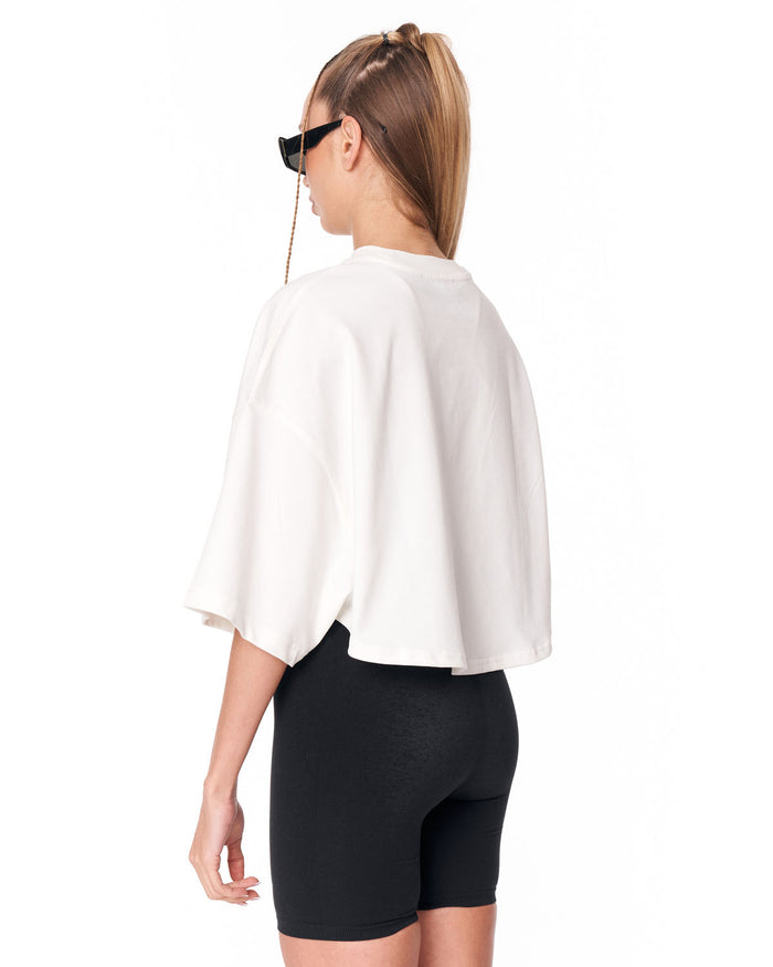 A woman shown from the back wearing a white cropped cape-style top and black fitted biker shorts against a plain white studio background.