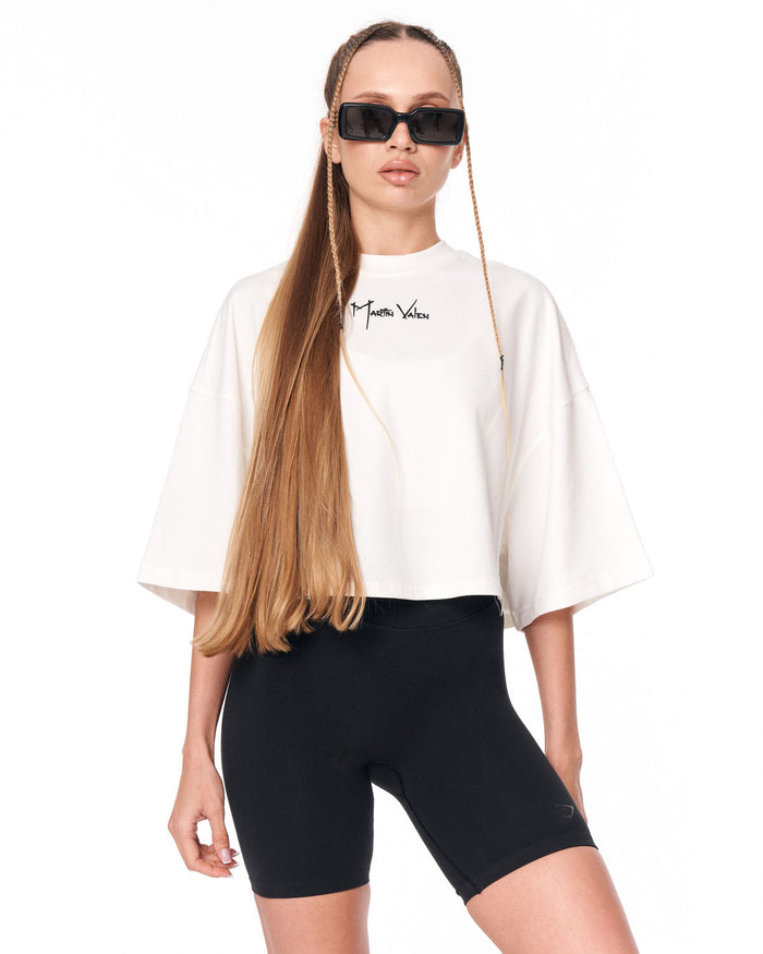 A model wearing a white cropped T-shirt with a black script logo on the chest, paired with black bike shorts and black sunglasses, against a plain white background.