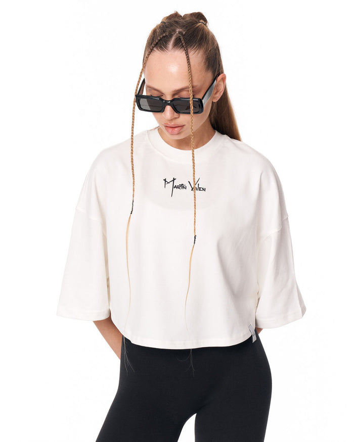 A woman wearing a white cropped t-shirt with a black script logo on the chest, paired with black pants and black sunglasses, set against a plain white background.