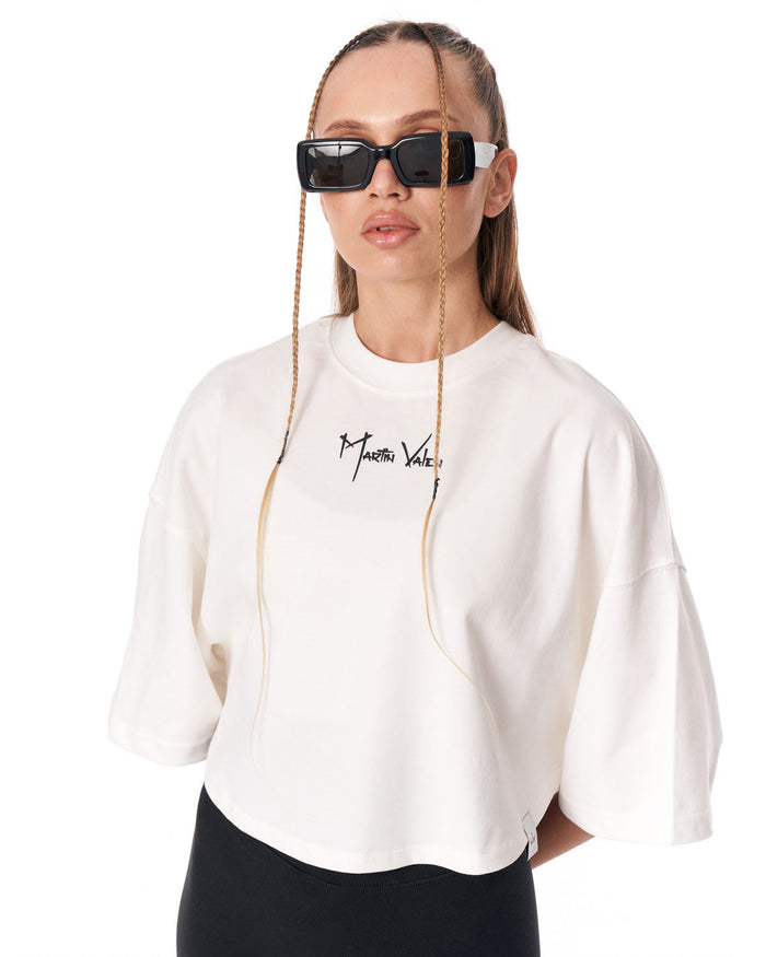 A model wearing a white cropped t-shirt with a black script logo on the chest, styled with black sunglasses and black bottoms against a plain white background.