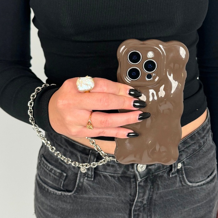 Phone with a shiny brown case and a silver chain strap, held by someone in black clothing.