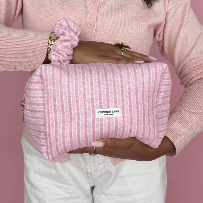 Pink-striped makeup bag and matching scrunchie set held by someone in a pink sweater, against a pink background.