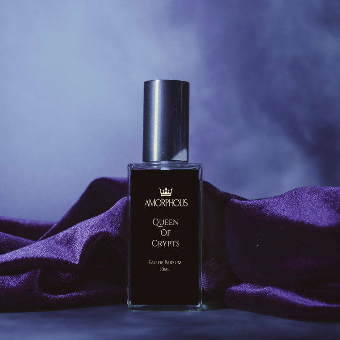 A bottle of Amorphous Queen of Crypts Eau de Parfum with a black label and silver cap, set against a smoky purple background with draped purple fabric.