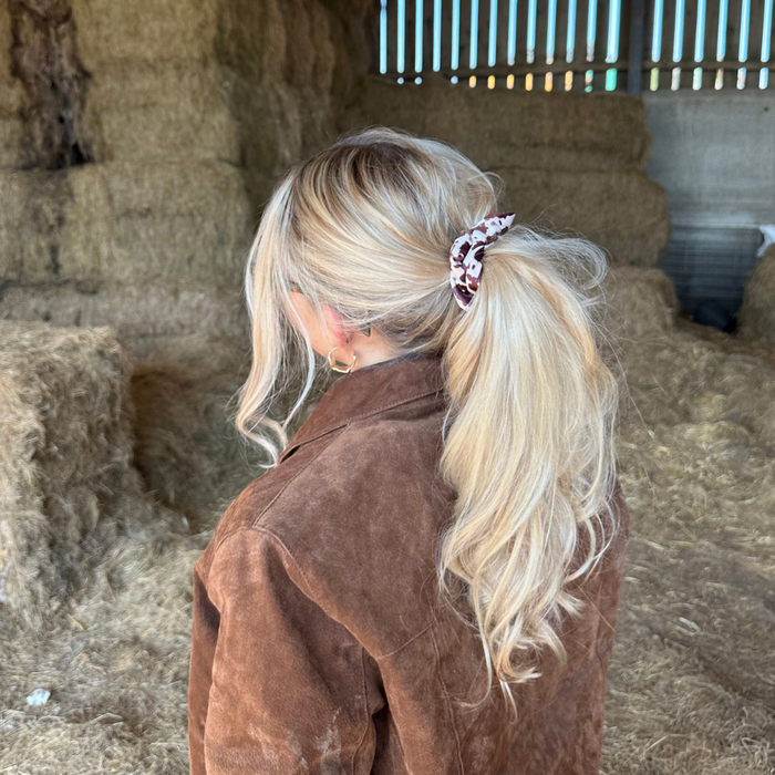 Woman with long blonde hair wearing a patterned Rodeo Cowgirl Scrunchie by Coconut Lane, standing in a barn with hay.