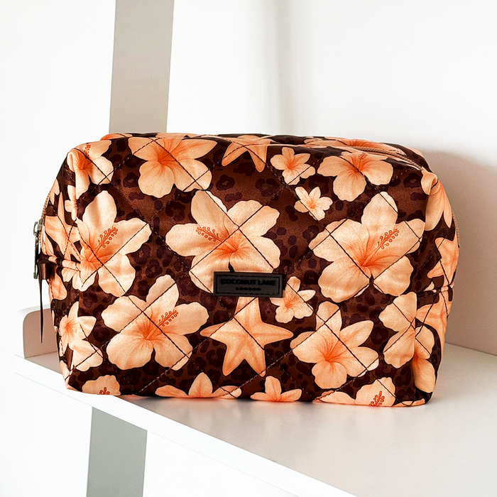 Satin quilted makeup bag with brown and orange floral pattern, zipped closed on a white shelf.