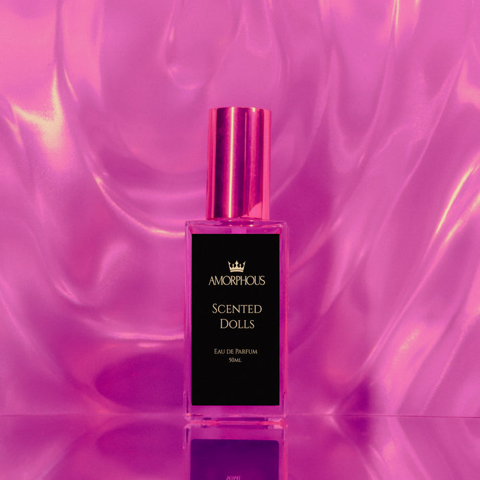 A clear rectangular perfume bottle with a shiny metallic pink cap and a black front label reading AMORPHOUS SCENTED DOLLS, centered against a reflective pink-purple satin-like background.