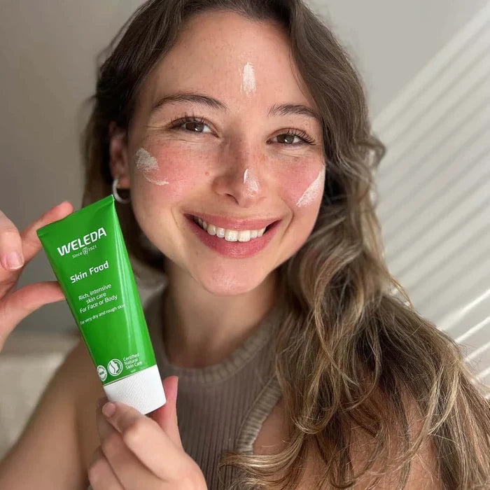 Smiling woman showing tube of Weleda skin food with swatches on her cheeks, forehead and nose.