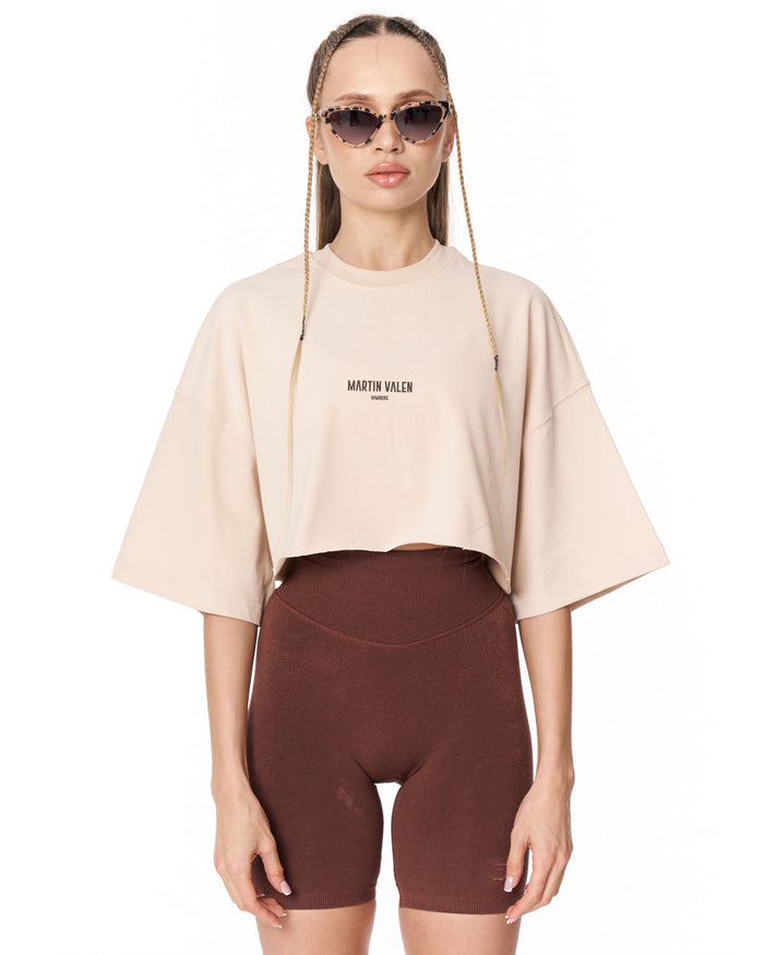 A model wearing a light beige cropped t-shirt with 'MARTIN VALEN' printed on the chest, paired with dark brown bike shorts, against a plain white background.