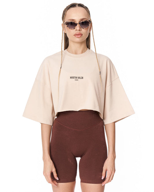 A model wearing a light beige cropped t-shirt with 'MARTIN VALEN' printed on the chest, paired with dark brown bike shorts, against a plain white background.