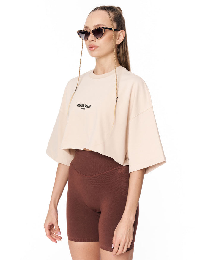 A woman modeling a cropped beige t-shirt with 'MARTIN VALEN' printed on the chest, paired with brown bike shorts, against a plain white background.
