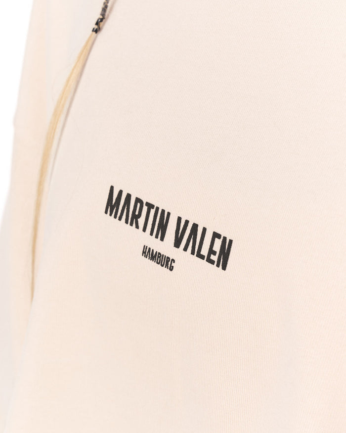 Close-up of a light beige or cream-colored garment with black printed text reading 'MARTIN VALEN' and a small portion of a zipper visible at the top left.