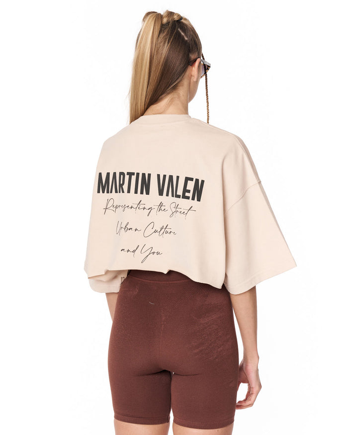 A model shown from the back wearing a light beige cropped t-shirt with 'MARTIN VALEN' printed in black across the back, paired with brown fitted shorts, against a plain white background.