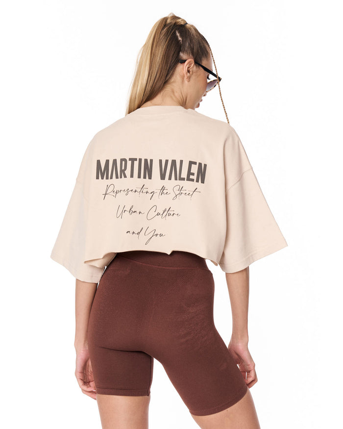 A woman shown from the back wearing a light beige cropped T-shirt with 'MARTIN VALEN' printed in dark lettering across the back, paired with brown fitted shorts against a plain white background.