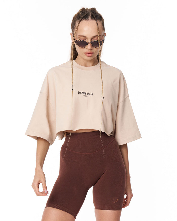 A model wearing a cropped beige t-shirt with 'MARTIN VALEN' printed on the chest and dark brown fitted biker shorts, set against a plain white background.