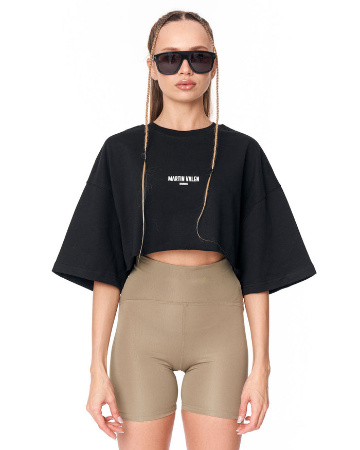 A model wearing a black cropped t-shirt with 'MARTIN VALEN' printed in white on the chest, paired with beige bike shorts, against a plain white background.