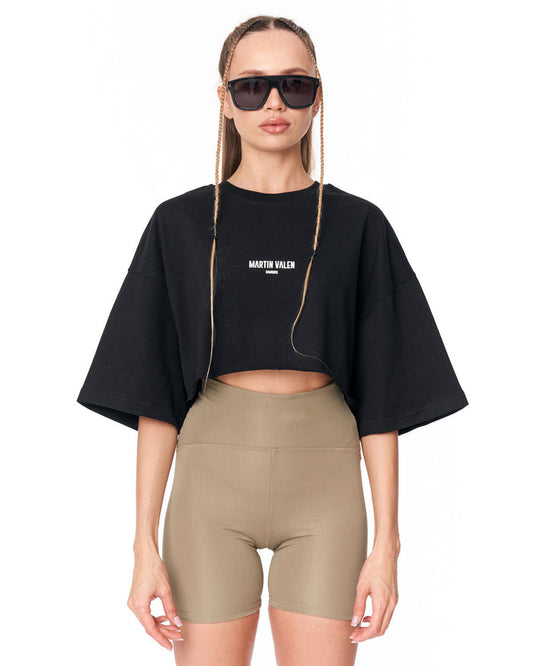 A model wearing a black cropped t-shirt with 'MARTIN VALEN' printed in white on the chest, paired with beige bike shorts, against a plain white background.