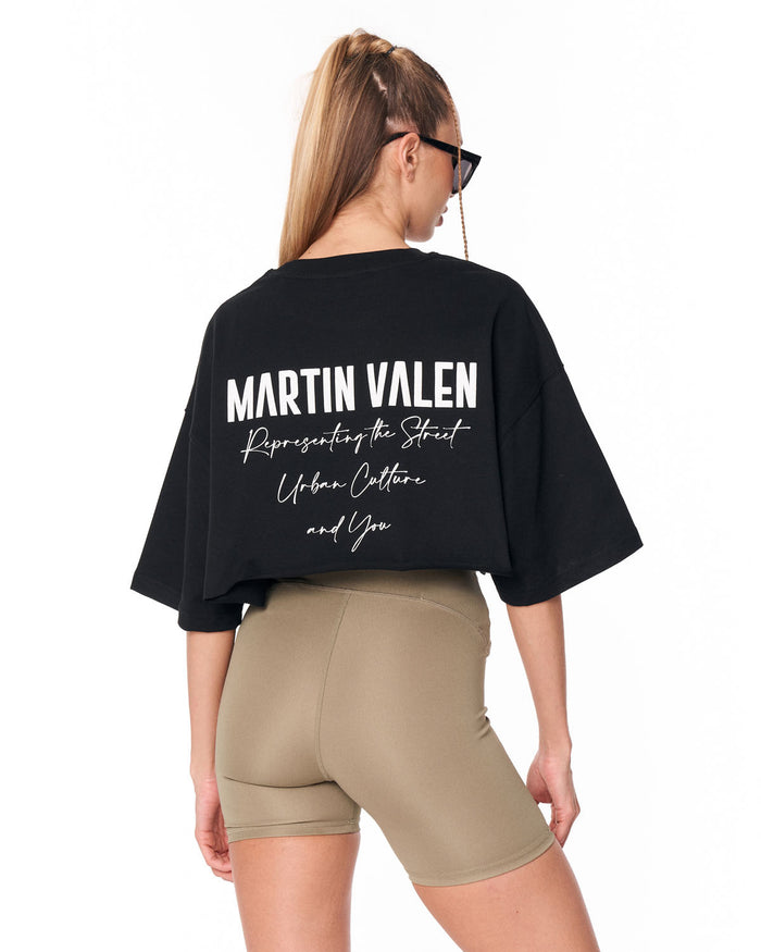 A woman shown from the back wearing a black oversized Martin Valen graphic T-shirt and beige fitted shorts against a plain white background.