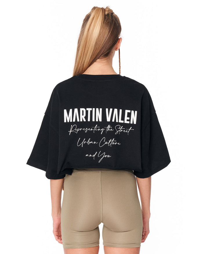 A woman shown from the back wearing a black Martin Valen graphic t-shirt and beige fitted shorts against a plain white background.