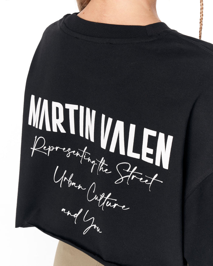 A close-up of a person wearing a black cropped T-shirt with large white text on the back reading 'MARTIN VALEN' and smaller script text below, against a plain white background.