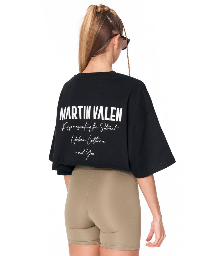 A woman shown from the back wearing a black oversized Martin Valen graphic t-shirt and beige fitted shorts against a plain white background.