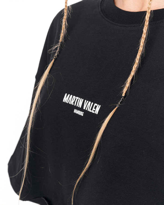 Close-up of a person wearing a black crewneck sweatshirt with white 'MARTIN VALEN' text printed on the chest, featuring blonde braided hair hanging down.