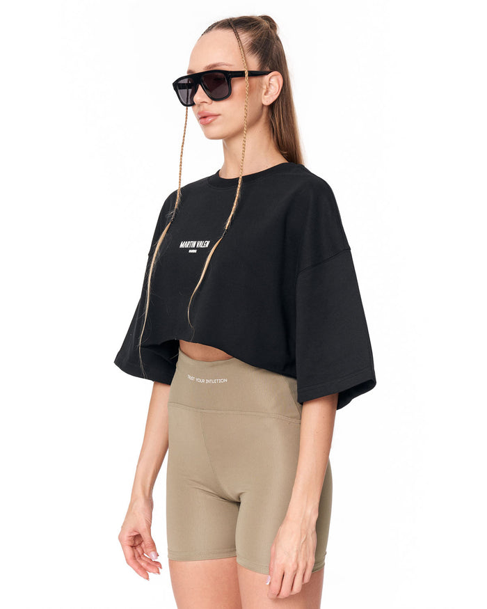 A woman modeling a black cropped t-shirt with a gold zipper detail and white text, paired with beige athletic shorts, against a plain white background.