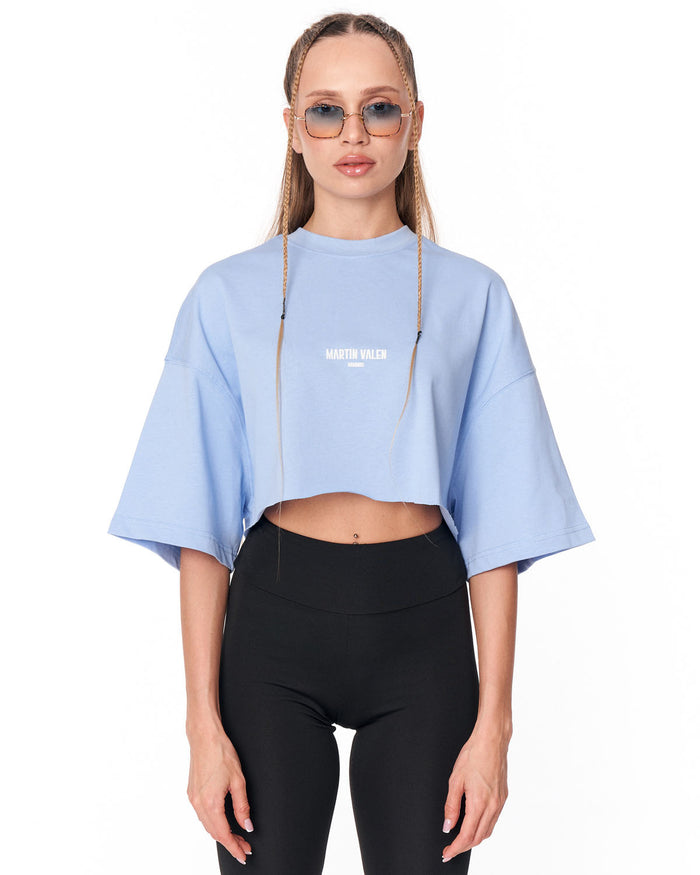A model wearing a light blue cropped t-shirt with 'MARTIN VALEN' printed in white on the chest, paired with black leggings, against a plain white background.