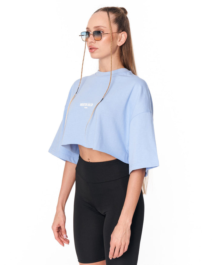 A woman modeling a light blue cropped t-shirt with a white chest graphic, paired with black biker shorts, against a plain white background.