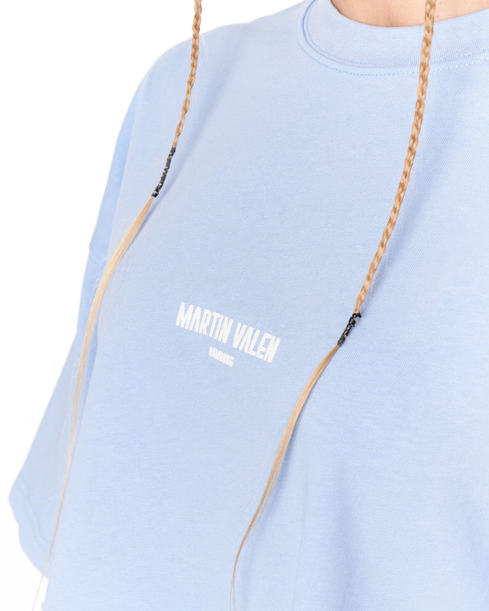 A close-up of a person wearing a light blue crewneck t-shirt with 'MARTIN VALEN' printed in white on the chest. The person has blonde braided hair hanging down the front of the shirt, set against a plain white background.