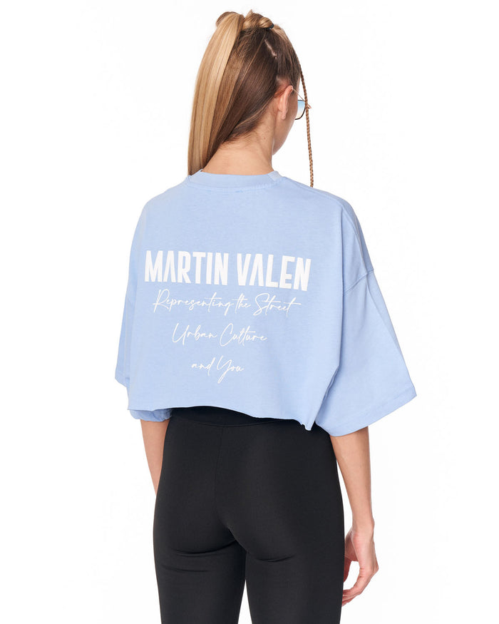 A woman shown from the back wearing a light blue oversized crop top with white text reading 'MARTIN VALEN' and smaller script text below, paired with black leggings, against a plain white background.