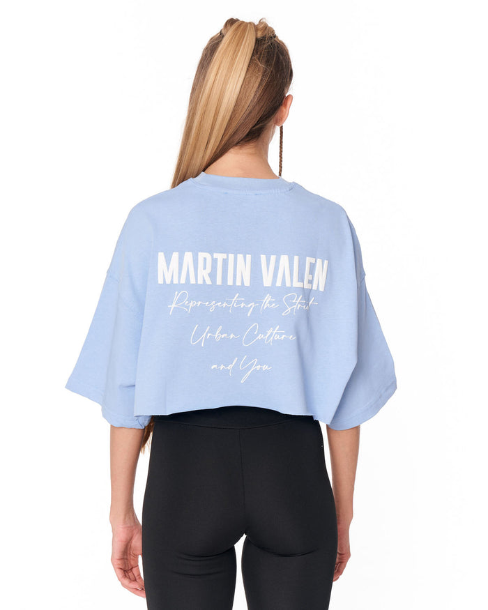 A woman viewed from the back wearing a light blue cropped T-shirt with white text reading 'MARTIN VALEN' and smaller script text below, paired with black leggings against a plain white background.