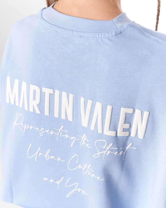 A close-up of a light blue crewneck sweatshirt featuring large white 'MARTIN VALEN' text and white script text on the front, worn by a model.