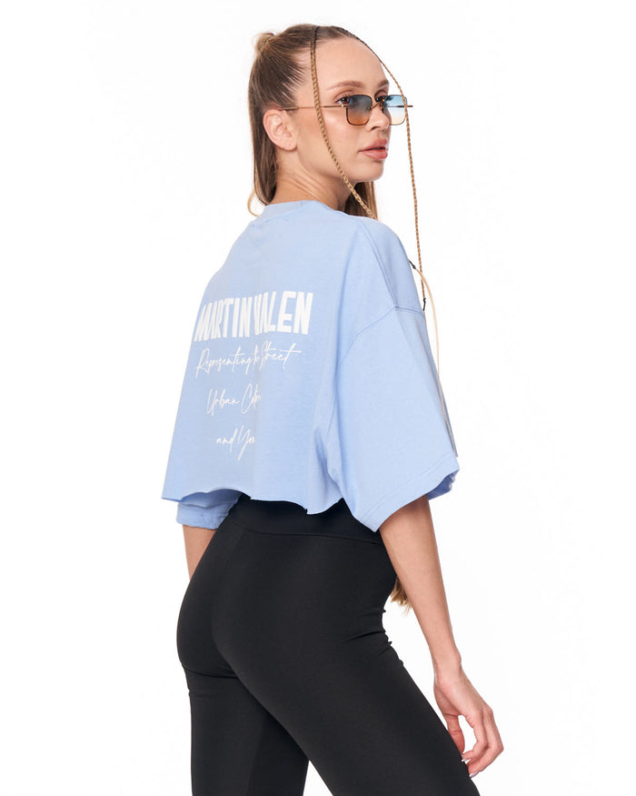 A woman modeling a light blue oversized cropped T-shirt from the back, featuring white text that reads 'MARTIN VALEN' and smaller script text below, paired with black leggings against a plain white background.