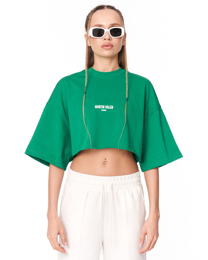 A model wearing a bright green cropped t-shirt with 'MARTIN VALEN' printed in white on the chest, paired with white sweatpants and white-framed sunglasses, against a plain white background.