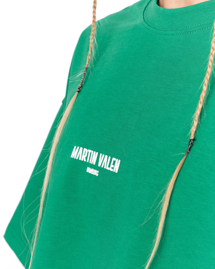 A close-up of a bright green crewneck t-shirt featuring the white printed text 'MARTIN VALEN' on the chest, worn by a model with blonde braided hair.
