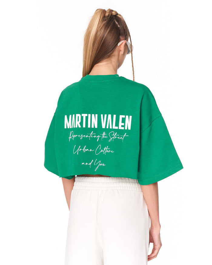 A model shown from the back wearing a bright green cropped T-shirt with 'MARTIN VALEN' printed in white across the back, paired with white pants, against a plain white background.