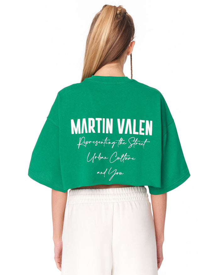 A woman shown from the back wearing a bright green cropped T-shirt with large white text reading 'MARTIN VALEN' and smaller script text below, paired with white pants against a plain white background.