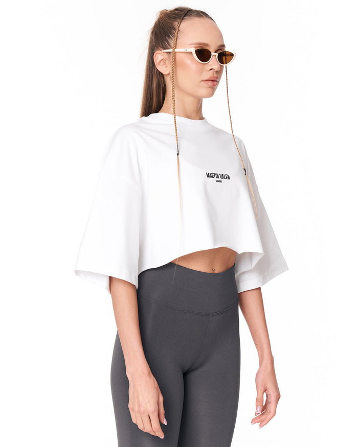 A model wearing a white cropped t-shirt with a black 'Martin Valen' logo on the chest, paired with dark gray leggings and gold-framed chain-link sunglasses, against a plain white background.