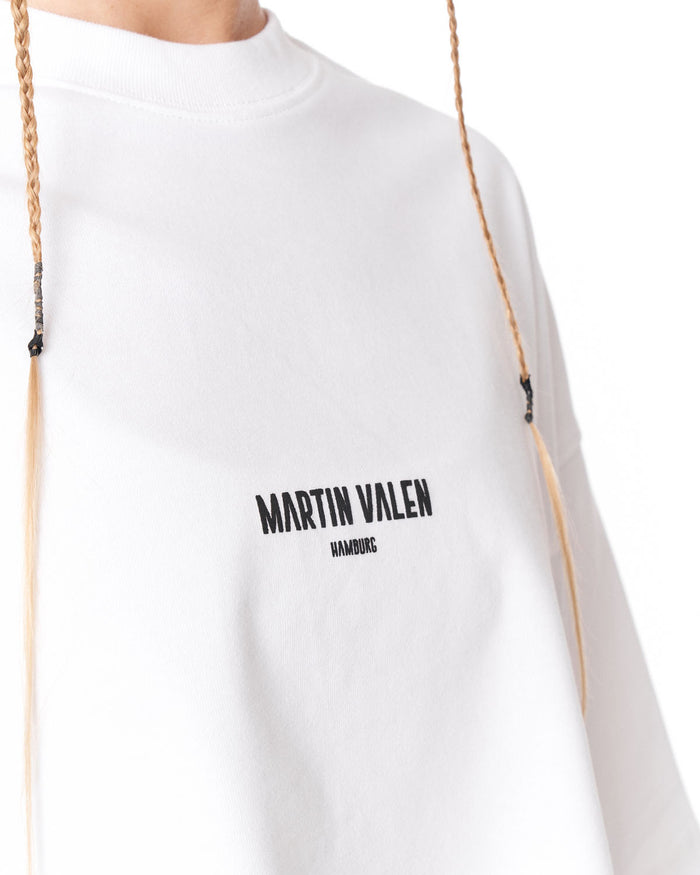 A close-up of a white crewneck sweatshirt featuring the black printed text 'MARTIN VALEN' and 'HAMBURG' on the chest, with braided drawstrings hanging down.