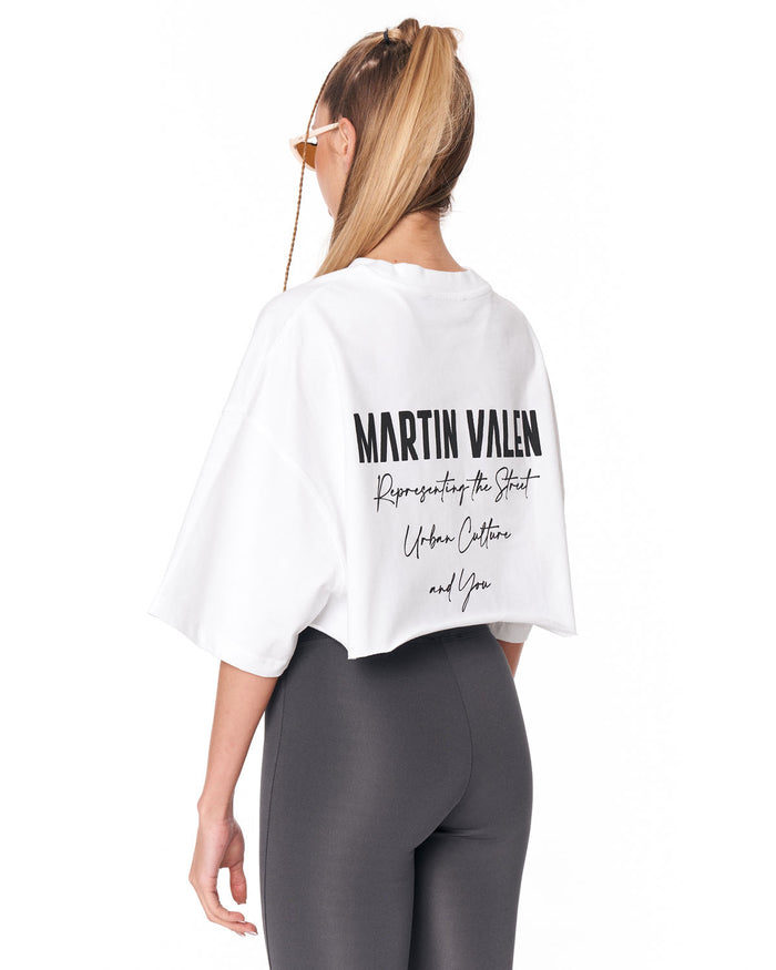 A woman shown from the back wearing a white cropped T-shirt with black Martin Valen text printed on the back, paired with dark gray leggings, against a plain white background.