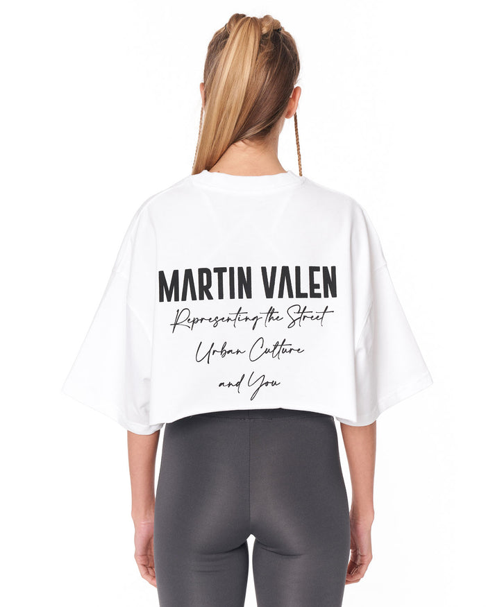 A woman shown from the back wearing a white cropped T-shirt with black Martin Valen text printed on the back, paired with dark gray leggings, against a plain white background.