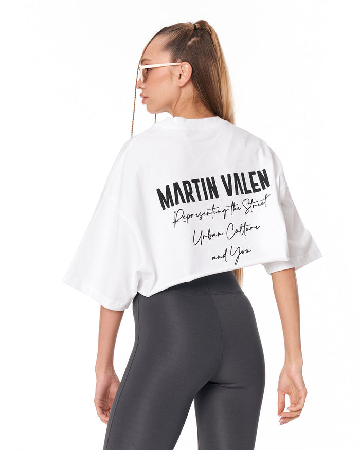 A woman shown from the back wearing a white cropped T-shirt with black Martin Valen text printed on the back, paired with dark gray leggings, against a plain white studio background.