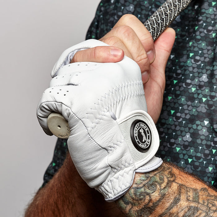 A close-up of a person gripping a golf club, wearing the Stealth Mode - Light - Glove for precision fit and durability. The cabretta leather glove sports a black and white circular logo on the wrist, with tattoos and a green patterned shirt visible.