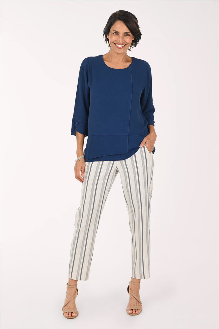 A woman modeling a navy blue long-sleeve top and white pants with thin gray vertical stripes, standing against a plain white studio background.