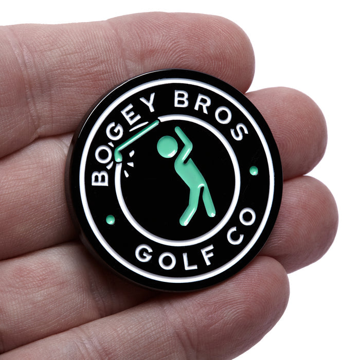 A hand holds The Big Coin - Ball Marker, a round black metal marker with "BOGEY BROS GOLF CO." in white text, a mint green stick figure swinging a club, all outlined by a white ring and two green dots.