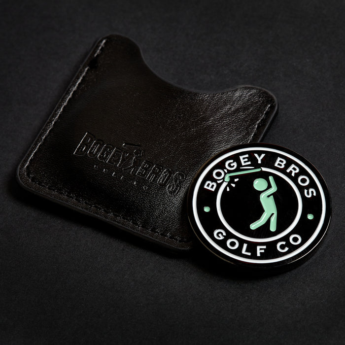 The Big Coin - Ball Marker, a circular marker with a golfer silhouette and double-sided design, is displayed next to a black leather case embossed with the brand name against a dark background.