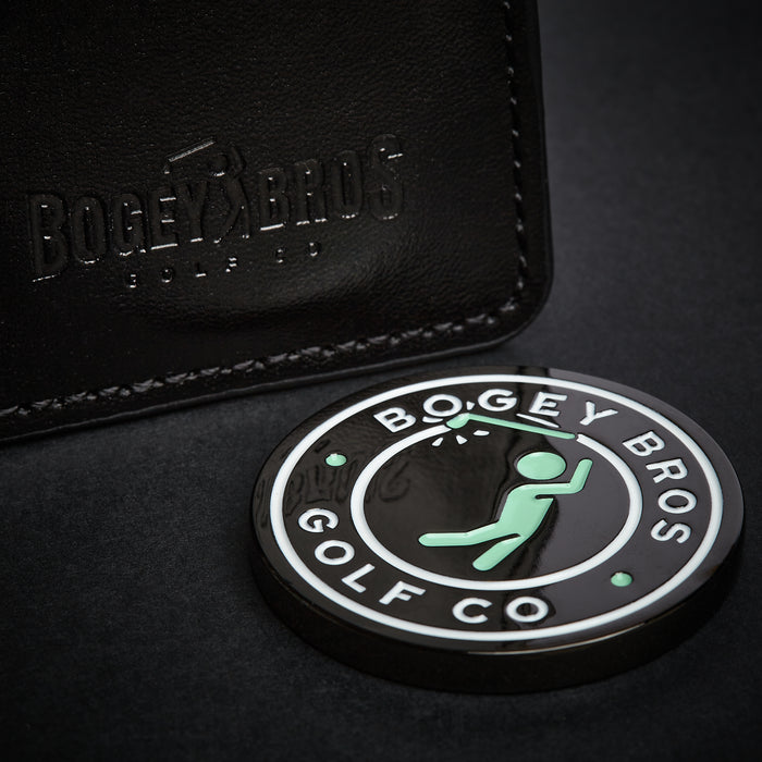 The Big Coin - Ball Marker, a round marker with a double face design featuring “BOGEY BROS GOLF CO” and a green stick figure swinging a club, rests on a dark surface beside black embossed Bogey Bros leather.